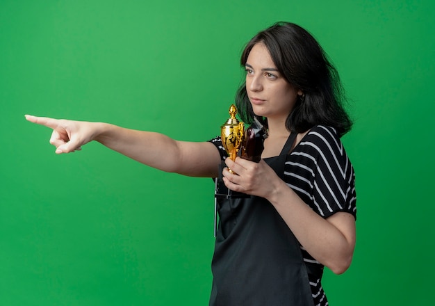 Belle jeune femme coiffeuse en tablier tenant le trophée et tondeuse pointant avec le doigt sur le côté avec une expression sceptique sur vert