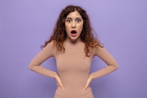 Belle jeune femme en col roulé beige étonnée et surprise avec les bras à la hanche debout sur le mur violet