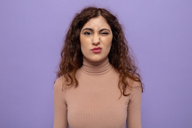 Belle jeune femme en col roulé beige faisant la bouche tordue avec une expression déçue debout sur violet