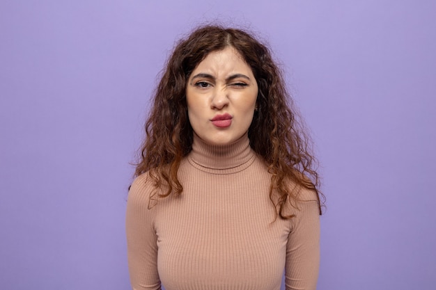 Belle jeune femme en col roulé beige avec un sourire sceptique sur le visage debout sur un mur violet