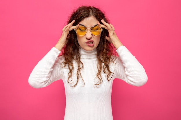 Belle jeune femme en col roulé blanc portant des lunettes jaunes à être mécontent de toucher ses tempes ayant des maux de tête