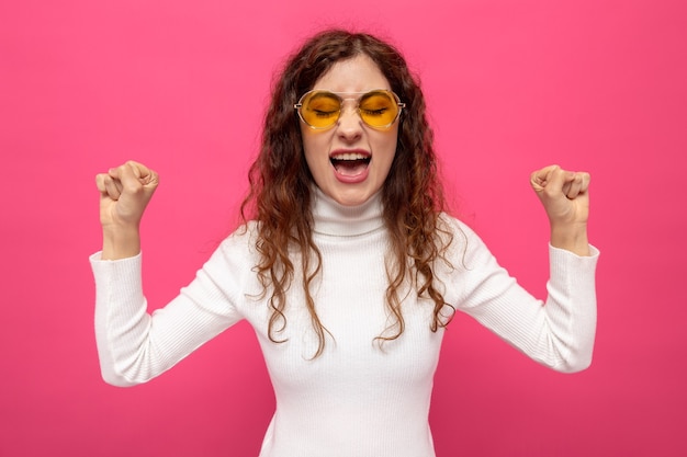 Belle jeune femme en col roulé blanc portant des lunettes jaunes levant les poings en criant avec une expression agacée debout sur rose