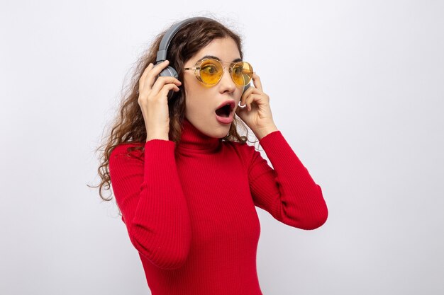 Belle jeune femme en col roulé rouge avec des écouteurs portant des lunettes jaunes regardant de côté étonné et surpris