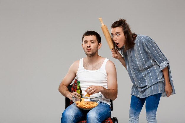 Belle jeune femme en colère contre un homme buvant de la bière, regardant le football sur un mur gris