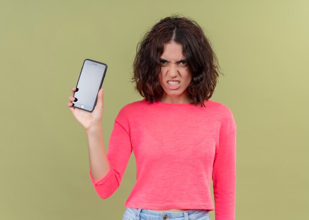 Belle jeune femme en colère tenant un téléphone mobile sur un mur vert isolé