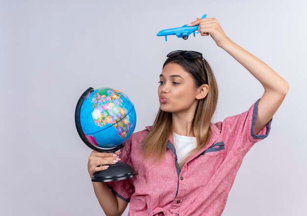 Une belle jeune femme confiante portant une chemise rouge à lunettes de soleil en regardant un globe tout en pilotant un avion jouet bleu sur un mur blanc