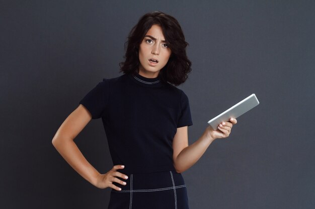 Belle jeune femme confuse posant sur un mur gris et tenant la tablette dans les mains