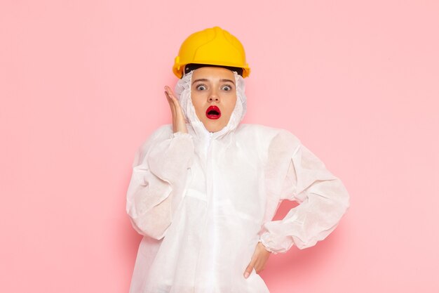 Belle jeune femme en costume blanc spécial et casque jaune posant avec une expression surprise sur rose