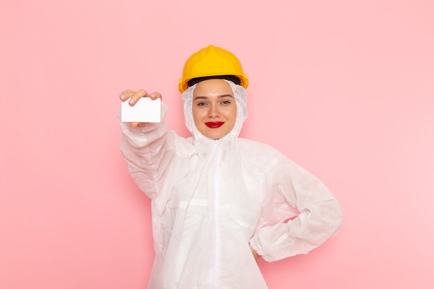 Belle jeune femme en costume blanc spécial et casque jaune tenant une carte blanche sur rose