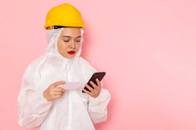 Belle jeune femme en costume blanc spécial et casque jaune tenant et utilisant le téléphone rose