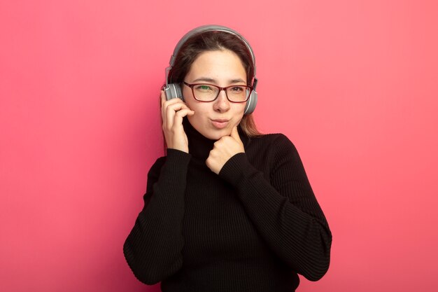 Belle jeune femme dans un col roulé noir et des lunettes avec des écouteurs à l'avant avec la main sur le menton heureux et positif debout sur le mur rose