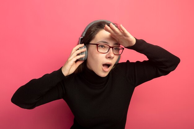 Belle jeune femme dans un col roulé noir et des lunettes avec des écouteurs à la recherche de loin avec la main sur la tête debout sur le mur rose