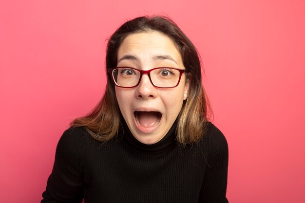 Belle jeune femme dans un col roulé noir et des lunettes hurlant d'être confus debout sur un mur rose