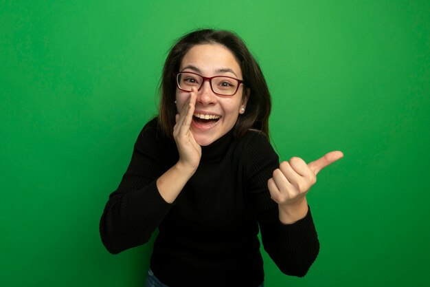 Belle jeune femme dans un col roulé noir et lunettes souriant chuchotant potins pointign avec le doigt sur le côté debout sur le mur vert