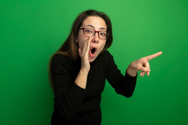 Photo gratuite belle jeune femme dans un col roulé noir et des lunettes à la surprise de chuchoter des potins pointign avec le doigt sur le côté debout sur le mur vert