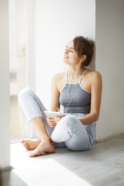 Belle jeune femme dans les écouteurs souriant regardant la fenêtre en appréciant la tenue de tablette en écoutant de la musique en streaming assis sur le sol sur le mur blanc