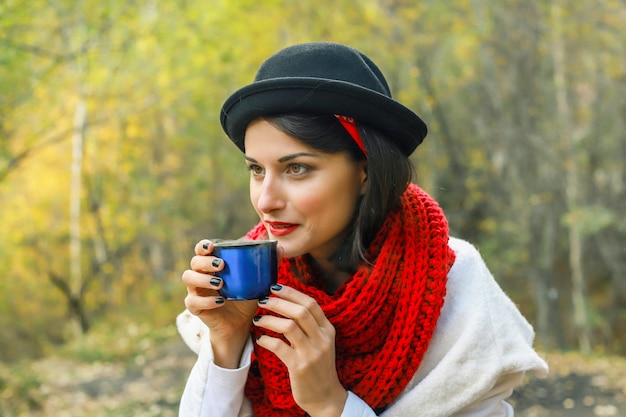 Belle jeune femme dans un manteau blanc au crochet écharpe rouge et chapeau noir tient une tasse dans ses mains