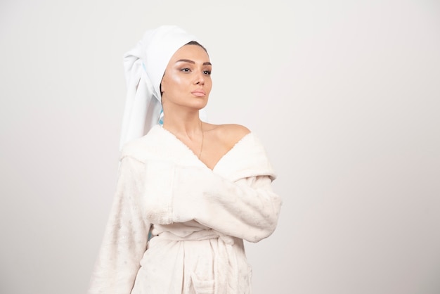 Belle jeune femme dans un peignoir blanc posant sur un mur blanc.