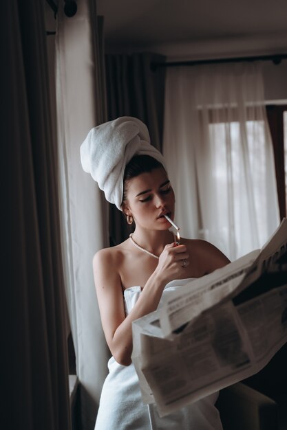 Belle jeune femme dans une serviette fume une cigarette et lit le journal