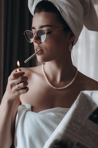 Belle jeune femme dans une serviette fume une cigarette et lit le journal