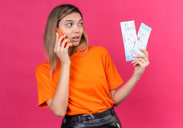 Photo gratuite une belle jeune femme dans un t-shirt orange parlant au téléphone mobile tout en tenant des billets d'avion sur un mur rose