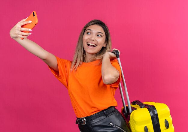 Une belle jeune femme dans un t-shirt orange souriant et prenant selfie avec téléphone mobile tout en tenant une valise jaune sur un mur rose