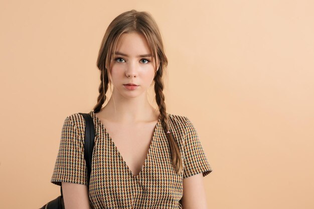 Belle jeune femme avec deux tresses en robe de tweed avec sac à dos noir sur l'épaule regardant pensivement à huis clos sur fond beige isolé