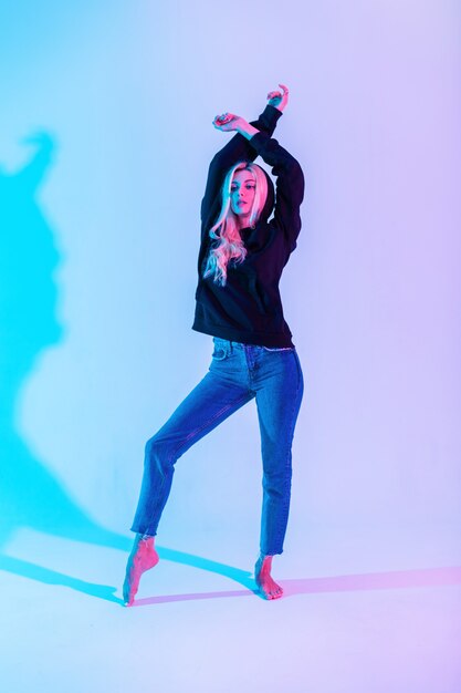 Belle Jeune Femme élégante Dans Un Sweat à Capuche Noir De Mode Avec Des Jeans Classiques Bleus Danse Sur Un Fond Clair Rose Néon Lumineux Photo Premium
