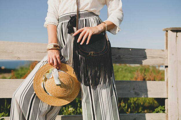 Belle jeune femme élégante, tendance de la mode printemps été, style boho, chapeau de paille, week-end de campagne, ensoleillé, souriant, amusant, lunettes de soleil, sac à main noir, pantalon rayé, détails, accessoires