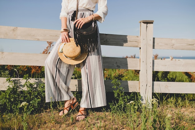 Belle jeune femme élégante, tendance de la mode printemps été, style boho, chapeau de paille, week-end de campagne, ensoleillé, souriant, amusant, lunettes de soleil, sac à main noir, pantalon rayé, détails, accessoires