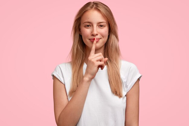 Belle jeune femme européenne joyeuse touche le doigt avant sur les lèvres, fait un geste de silence