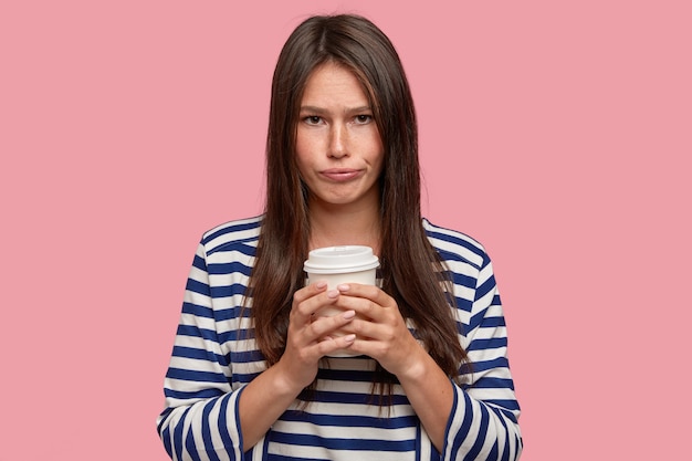 Belle jeune femme a une expression triste et misérable, tient un gobelet en papier jetable, boit du café, se sent bouleversée