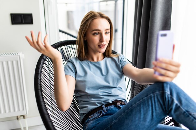 Belle jeune femme faisant du selfie par son téléphone intelligent et souriante assise sur une grande chaise à la maison