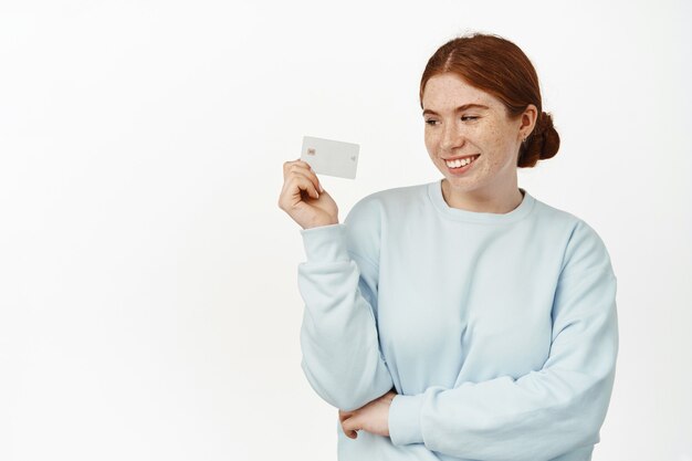 Belle jeune femme, fille aux cheveux rouges tenant une carte de réduction de crédit et souriante, l'air heureuse, payant en magasin sans contact, debout sans soucis sur blanc