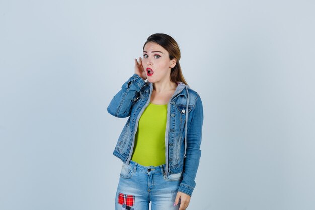 Belle jeune femme gardant la main derrière l'oreille tout en entendant un secret en tenue de denim et l'air surprise. vue de face.