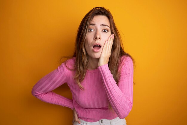 Belle jeune femme en haut rose regardant la caméra étonnée et surprise debout sur fond orange