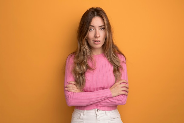 Belle jeune femme en haut rose regardant la caméra avec une expression sérieuse et confiante debout sur fond orange