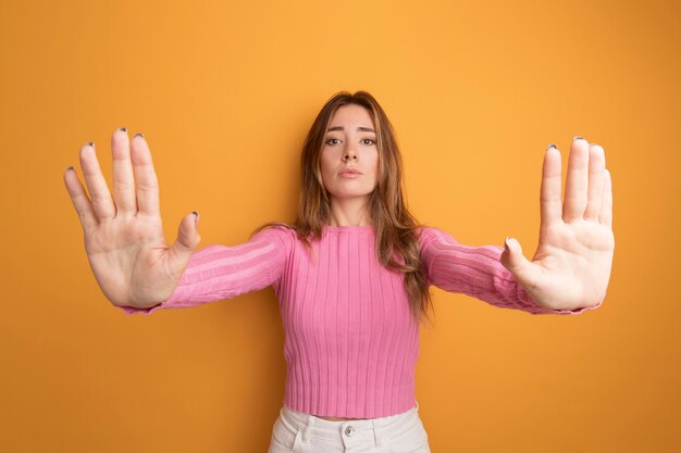 Belle jeune femme en haut rose regardant la caméra avec un visage sérieux faisant un geste d'arrêt avec les mains debout sur fond orange