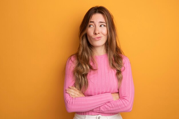 Belle jeune femme en haut rose regardant de côté faisant la bouche tordue avec une expression déçue debout sur orange