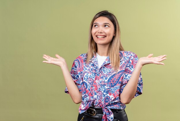 Une belle jeune femme heureuse portant une chemise imprimée paisley souriant et ouvrant les mains tout en regardant sur un mur vert