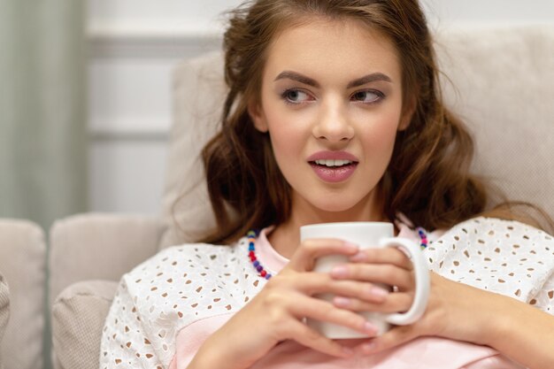 Belle jeune femme joyeuse tenant une tasse de café et regardant ailleurs avec le sourire à la cuisine à la maison