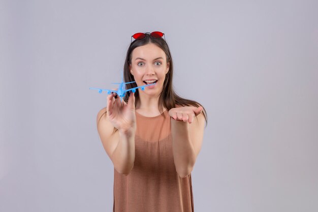 Belle jeune femme avec des lunettes de soleil rouges sur la tête tenant un avion jouet pointant avec le bras de la main à la caméra souriant avec un visage heureux sur un mur blanc