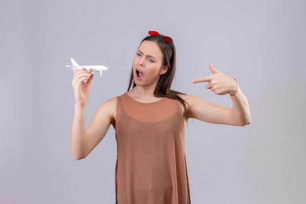 Belle jeune femme avec des lunettes de soleil rouges sur la tête tenant un avion jouet pointant avec le doigt sur elle surpris et étonné sur le mur blanc