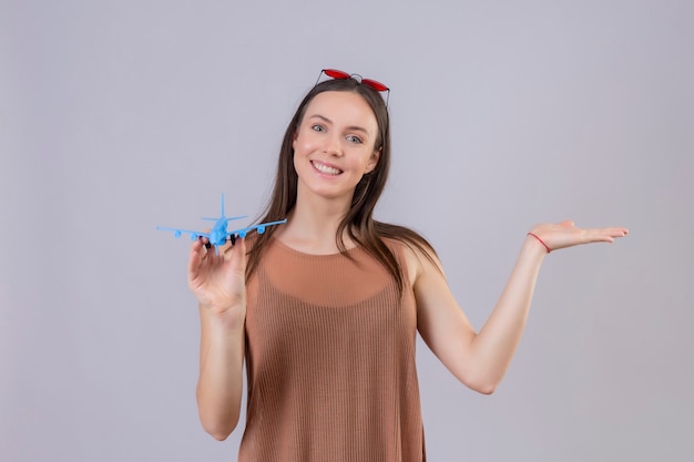 Belle jeune femme avec des lunettes de soleil rouges sur la tête tenant un avion jouet présentant le bras de la main souriant avec un visage heureux debout sur fond blanc