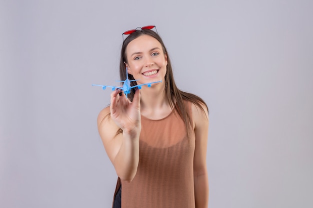 Belle jeune femme avec des lunettes de soleil rouges sur la tête tenant un avion jouet souriant avec un visage heureux sur un mur blanc