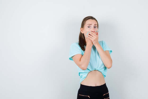 Belle jeune femme avec les mains sur la bouche en t-shirt, pantalon et l'air effrayé. vue de face.