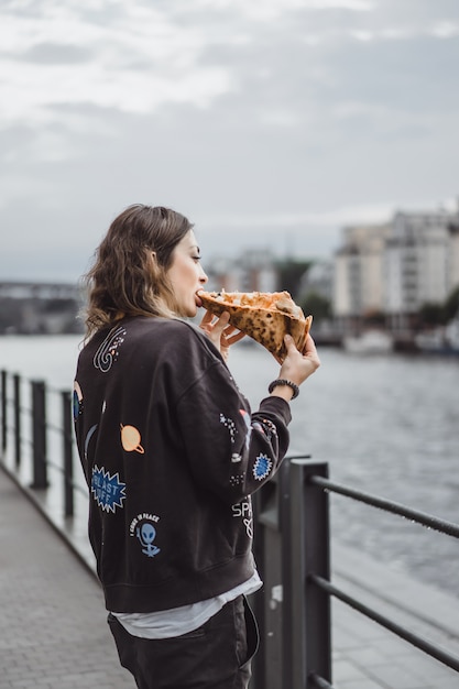 Belle jeune femme mangeant une part de pizza dans la rue