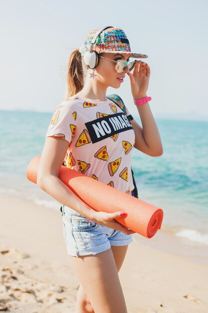 Belle jeune femme marchant sur la plage avec tapis de yoga, écouter de la musique sur les écouteurs, style swag sport hipster, short en jean, t-shirt, sac à dos, casquette, lunettes de soleil, ensoleillé, week-end d'été, joyeux