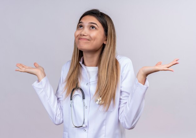 Belle jeune femme médecin portant blouse blanche avec stéthoscope à la confusion et incertain en haussant les épaules n'ayant pas de réponse