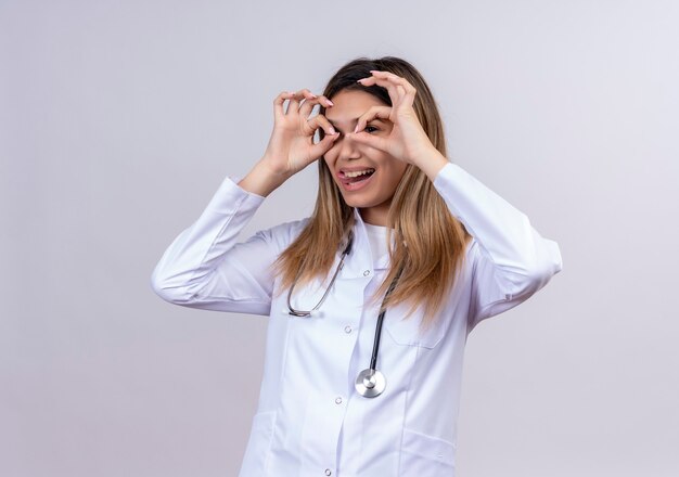 Belle jeune femme médecin portant blouse blanche avec stéthoscope faisant des signes ok avec les doigts comme des jumelles regardant à travers les doigts qui sort la langue